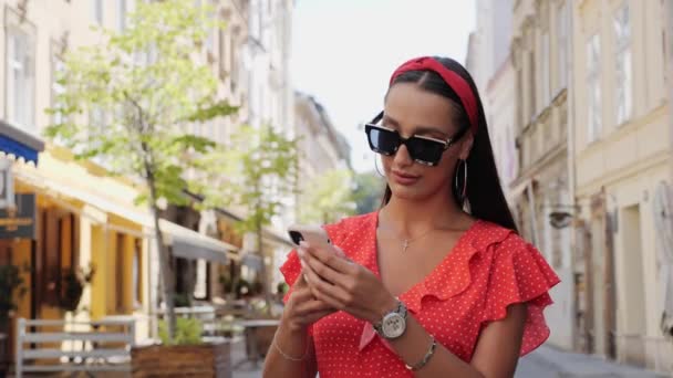 Stylish and beautiful tourist in sunglasses walks down the city street, where there are many cafes. The girl uses her phone to view the sights of the city. — Stock Video