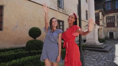 İki turist arkadaş kameranın önünde poz veriyor ve şehir sokaklarında el sallıyorlar..