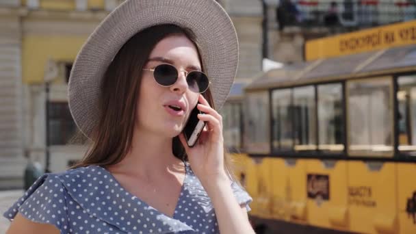 Стильна і красива дівчина в сонцезахисних окулярах і капелюсі говорить на мобільному телефоні на фоні старого міста, повного людей . — стокове відео