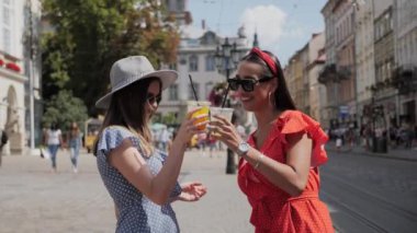 İki kız limonata içiyor ve insanlarla dolu bir caddede gülümsüyor..
