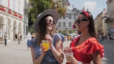 İki çekici kız limonata içiyor ve insanlarla ve kafelerle dolu bir caddede gülümsüyor..