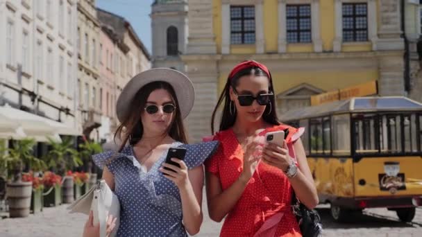 Twee vrolijke stijlvolle en vriendelijke meisjes in een zonnebril wandelen door de stad en het gebruik van de smartphone voor likes, volgers, opmerkingen voor sociale media. — Stockvideo