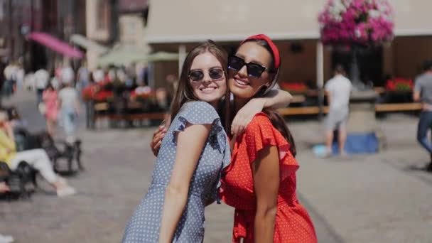 Duas namoradas bonitos elegantes em belos vestidos de verão andando pela cidade urbana, rindo, abraçando e se divertindo. A rua está cheia de cafés e pessoas. . — Vídeo de Stock