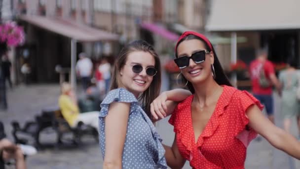 Twee stijlvolle, aantrekkelijke beste vrienden in mooie zomerjurken die door de stad wandelen, lachen, knuffelen en plezier hebben. De straat is vol met cafés en mensen. — Stockvideo