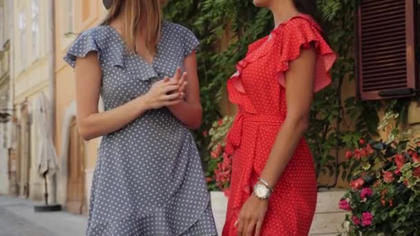 Yazlık elbiseli ve güneş gözlüklü iki arkadaş şehirde çiçekli güzel bir evin arka planında konuşup gülüyorlar.. — Stok video