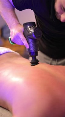 Siyahlar giymiş yetenekli erkek terapist müşterilerine silahla perküsyon masajı yapıyor. Müşteri, klinik ortamında kas iyileşmesine yardım ederek derin doku desteğini alır..