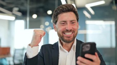 Heyecanlı bir iş adamı, takım elbise giymiş, zaferi bir yumrukla kutlarken modern bir ofiste akıllı telefondan olumlu haberler okuyor. Coşkulu bir profesyonel büyük başarı elde etti..