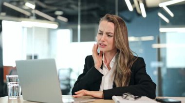 Siyah takım elbiseli hasta iş kadını modern bir ofiste şiddetli diş ağrısı çekiyor. Rahatsızlıktan bunalan yönetici, dizüstü bilgisayarla çalışırken sıkıntılı ve acı dolu bir ifade sergiliyor..