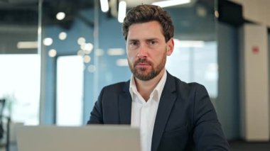 Koyu takım elbiseli ve beyaz gömlekli gülümseyen işadamı modern bir ofiste dizüstü bilgisayarda çalışır, sonra yukarı bakar ve neşeyle gülümser, başarıyı ve güveni iletir..