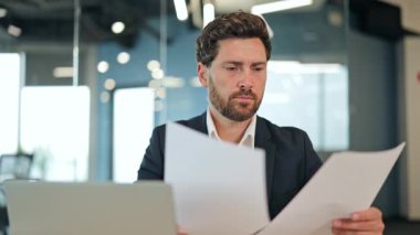 Takım elbiseli ciddi bir işadamı modern ofis masasında özenle evrak ve dizüstü bilgisayar üzerinde çalışıyor, ciddi bir konsantrasyon ve görevlere odaklandığını gösteriyor..