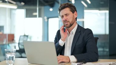 Takım elbiseli ve beyaz gömlekli sıkıntılı işadamı, modern bir ofiste şiddetli diş ağrısı ya da çene ağrısı çekiyor. Erkek, rahatsızlığı ve sağlık sorununu gösteren bir dizüstü bilgisayar üzerinde çalışıyor..