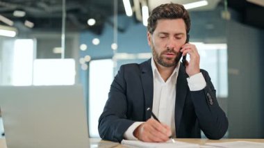 İş adamı modern ofis masasında önemli bir telefon görüşmesi yapıyor. Koyu renk takım elbiseli başarılı erkek verimliliği ifade eden notlar yazar. Neşeli adam iş ortamında konuşmaktan hoşlanır..