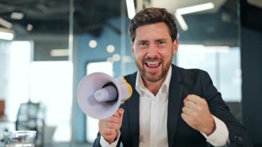 Takım elbiseli ve beyaz gömlekli enerjik işadamı modern bir ofiste megafona bağırıyor, heyecan ve kararlılığını ifade ederken aynı zamanda işaret ediyor..