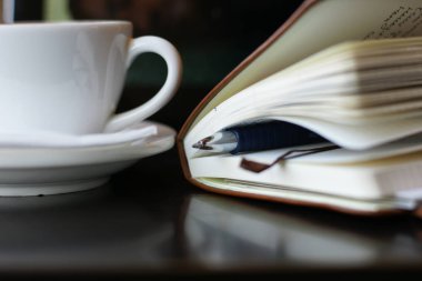 Bir fincan kahve ve masanın üzerinde kalem olan bir defter. 