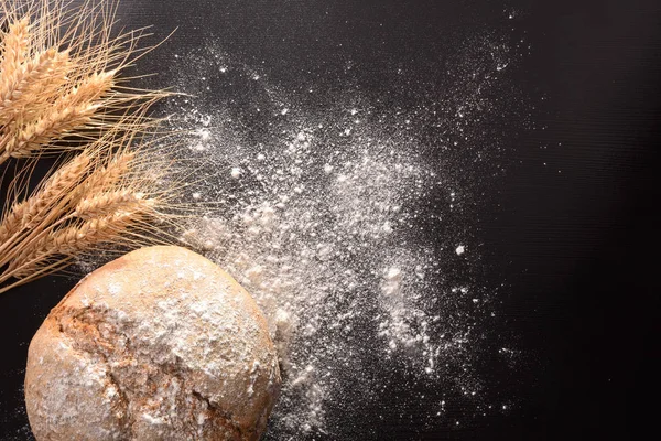 Loaf of bread with flour and wheat on black wooden table close up. Top view. Horizontal composition