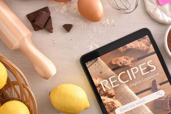 Tablet with online recipes app and pastry ingredients background. Use of the digital devices to cook. Concept of recipes in digital book. Horizontal composition. Top view
