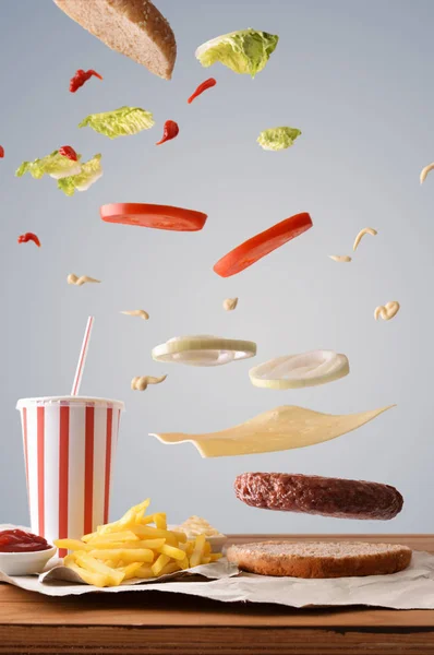 Creative breakdown of ingredients of a burger on wooden table and chips and refreshment accompaniment with gray background. Front view. Vertical composition.