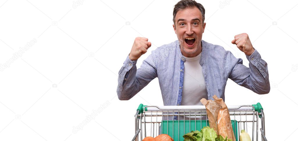 Happy supermarket customer raising hands with clenched fists with gesture of success with shopping cart full of food isolated white
