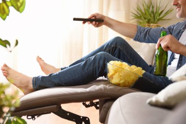 Kanepede rahatça televizyon seyreden adam elinde uzaktan kumandayla birayla atıştırıyor..