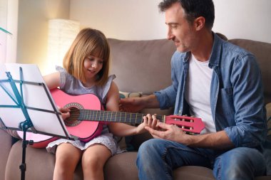 Öğretmen elinde pembe gitarıyla oturma odasındaki koltukta oturan bir kıza ders veriyor.