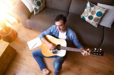 Gitar besteleyen bir adam evdeki oturma odasında yerde oturuyor. Üst görünüm.
