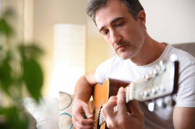 Evdeki oturma odasında beyaz kısa kollu akustik gitar çalan adamın ayrıntıları.