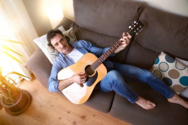 Rahatlamış ve gülümseyen bir adam evdeki bir odada gitar çalıyor. Üst görünüm.