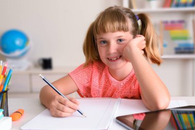 Evde ders çalışan mutlu bir kız. Masasında tablet olan bir not defterine yazıyor.