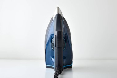 Mavi buhar ütüsü, beyaz bir masanın üzerinde dikey olarak duruyor. Kabzanın sapını ve ısı seçicisini detaylıca gösteriyor. İzole edilmiş beyaz bir arkaplanı var. Ön görünüm.