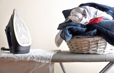 Ütü masasında dikey duran buharlı ütüyle giysileri ütüleme kavramı ve bir sepet dolusu giysi. Ön görünüm.
