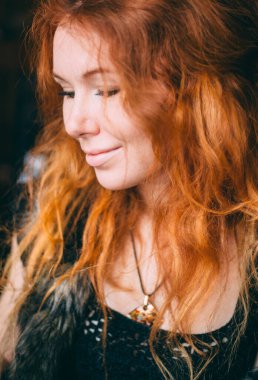 Makyajlı, siyah bir elbise ve yapay kürkten bir yelek giymiş güzel kızıl saçlı, kıvırcık bir kızın portresi..