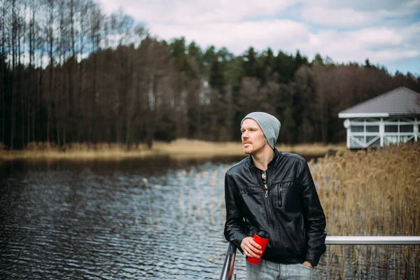 Pensive man in a leather jacket and hat stands and thinks outdoors. Good looking male model looking pensive at lake.