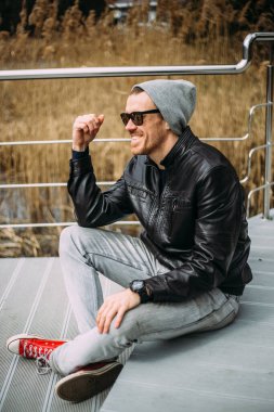 Deri ceketli ve şapkalı olumlu bir adam ayakta ve dışarıda gülümsüyor. Yakışıklı erkek model kamerada eğleniyor..