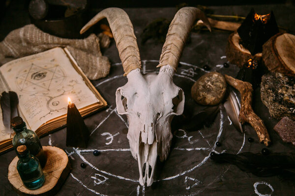 White goat scull with horns, open old book (text - untranslatable, fictional language), black candles on witch table. Occult, esoteric, divination and wicca concept. Halloween, Day of the dead concept