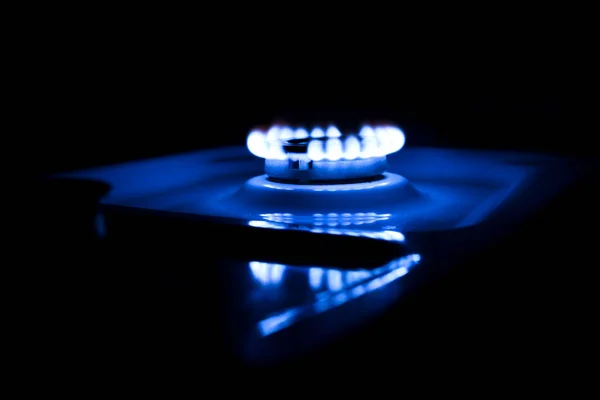 Blue flame from a burner in a gas stove on. Illuminated flame on a dark background, photo taken at night. A slight reflection in the white casing of the gas stove.