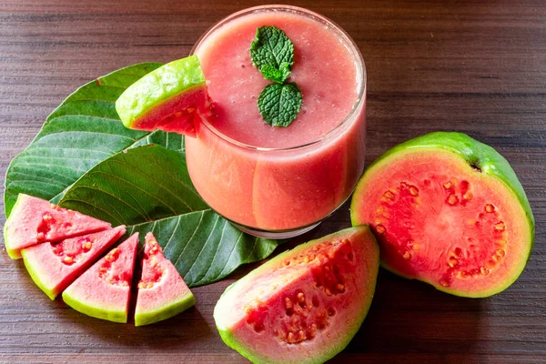 Delicious red guava juice alongside a slice of guava, in the background guavas and leaves on rustic wood.