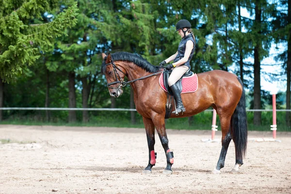 Training process. Young teenage girl riding bay horse on arena at equestrian school. Colored outdoors horizontal summertime image.