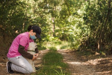 Coronavirus salgını sırasında parkta kaniş köpeğiyle oynayan tek kullanımlık tıbbi maske takan bir kadın. Hayvan yürüyüşü.