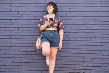 Beyaz kadın yanlış bir maske takıyor ve elinde akıllı telefonuyla gülümsüyor. Coronavirus önleme kavramı.