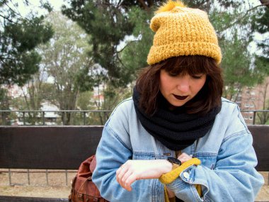 Sarı yünlü şapkalı genç bir kadın bankta oturmuş saatini kontrol ediyor. Sonbahar şehir konsepti.