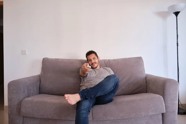 Portrait of a relaxed young man sitting on a sofa watching television and changing channels with the remote control on his hands.