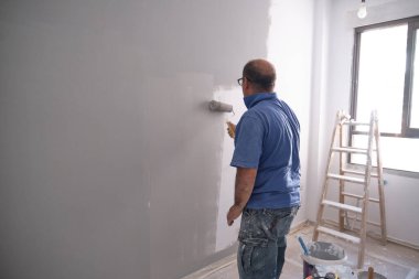 Gerçek bir son sınıf boyacısı gri bir duvarı boya rulosuyla boyuyor. Çok pis bir işçiydi. Boyacı mesleği kavramı.