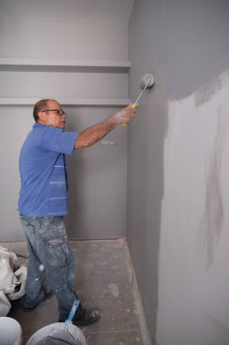 Gerçek bir son sınıf boyacısı gri bir duvarı boya rulosuyla boyuyor. Çok pis bir işçiydi. Boyacı mesleği kavramı.