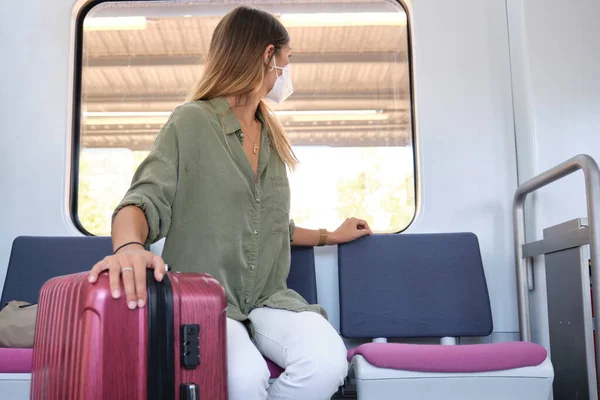 Young woman wearing face mask and carrying a suitcase looking through the window of the train. New normal travel concept.