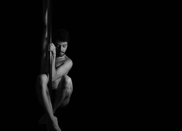 Dramatic dark portrait of a young man hanging from a pole dance bar with his eyes closed. Dark background studio shot. Black and white image.