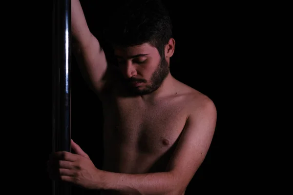 Dramatic dark portrait of a young man with a pole dance bar. Dark background studio shot.