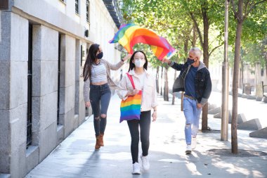 Yüzü maskeli üç arkadaş grubu sokakta LGBT bayrağı sallıyor. Salgın zamanlarında LGBT gurur kutlaması.