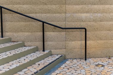 Bir dizi gri basamak, parlak siyah korkulukların yer aldığı bir binanın girişine çıkıyor. Dokulu duvar, merdivenlerin pürüzsüz yüzeyi ile çelişiyor ve modern bir estetik yaratıyor..