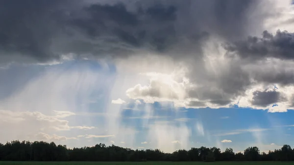 Misty rain and dark clouds in blue sky over field and trees