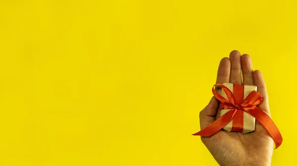 banner In hand, a handmade gift box is decorated with a red bow, wrapping paper on a yellow background. Holiday concept. space for text.the view from the top. copy space. the view from the top.